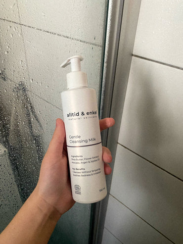 Hand holding a bottle of Gentle Cleansing Milk in front of a shower.